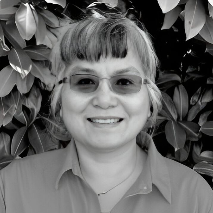 Smiling person with glasses, leafy background.