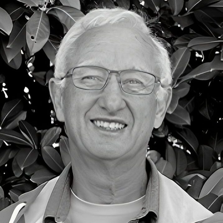 Smiling man with glasses, leafy background.